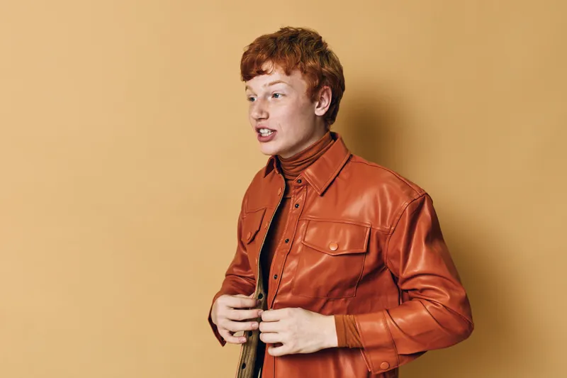Young man with red hair wearing orange leather jacket and turtleneck standing against beige background in casual fashion style portrait.