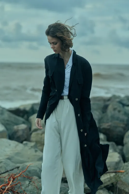 Woman in a long coat and white trousers walks along windy coastal rocks, sea in background, moody fashion scene with natural lighting