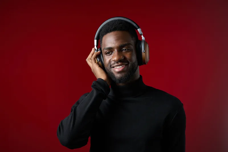 Young African man wearing headphones, smiling joyfully against a vibrant red background, showcasing a modern and stylish look in a cozy black turtleneck