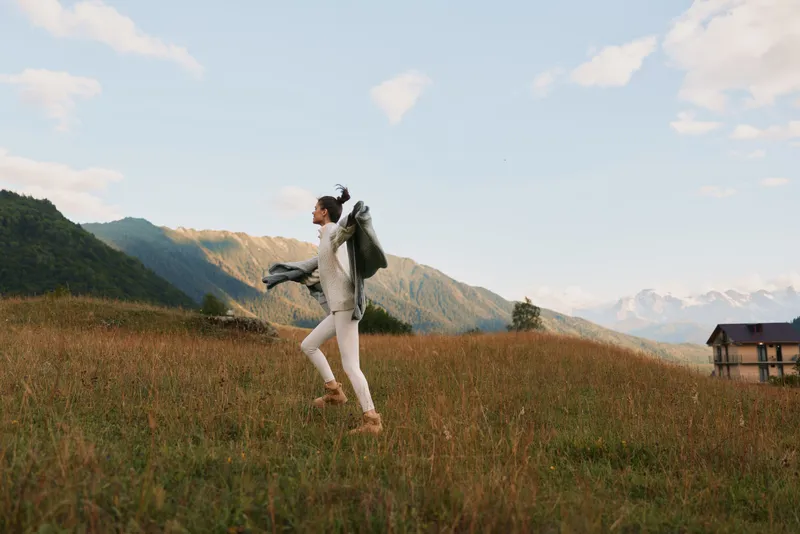 Young woman with backpack hikes across a grassy field toward distant mountains, radiating carefree adventure and energy in an open landscape at golden hour with soft breeze.