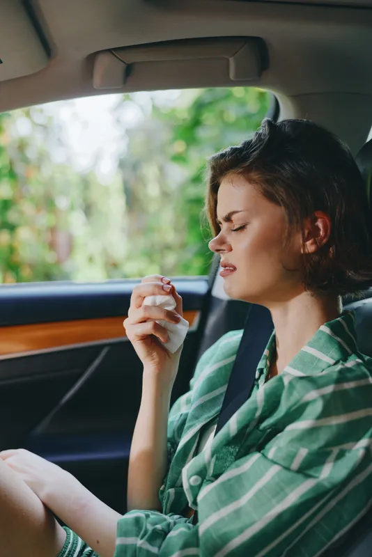 A woman sits in a car, looking unwell and holding a tissue, captured in natural light.