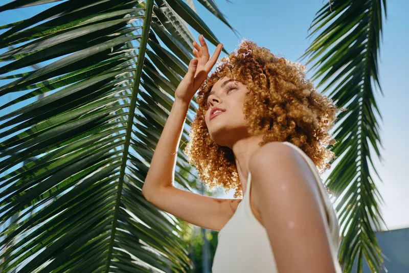 young woman with curly hair enjoying sunlight under palm leaves outdoors, clear blue sky, summer vibe, natural beauty and relaxed mood captured
