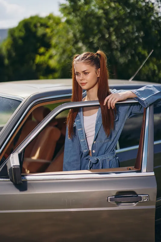 Young woman with long ponytail in denim outfit leaning against a vintage car, showcasing a vibrant outdoor setting and exuding a carefree summer vibe.