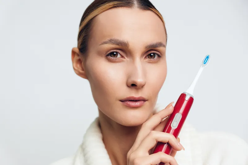Young woman holding red electric toothbrush close to face, neutral expression, clear skin, white background, personal care, dental hygiene concept, close-up portrait, clean modern look