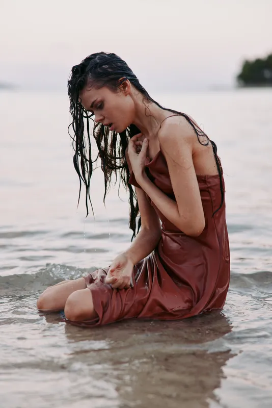 Young woman in a wet dress kneeling in shallow water with wet hair, expressing calm and introspection near the shore during soft natural light.