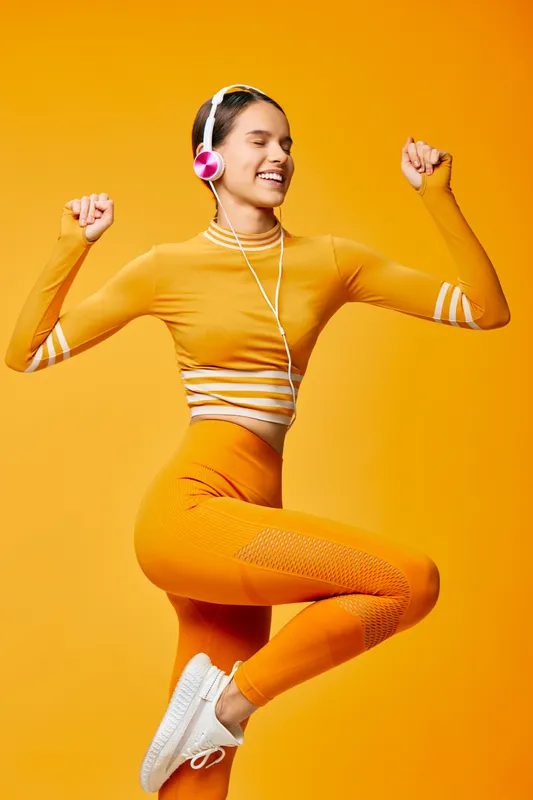 Fitness, sport, woman in bright orange athleisure dances mid-air with headphones, showcasing athletic energy and vibrant movement against a bold studio backdrop.