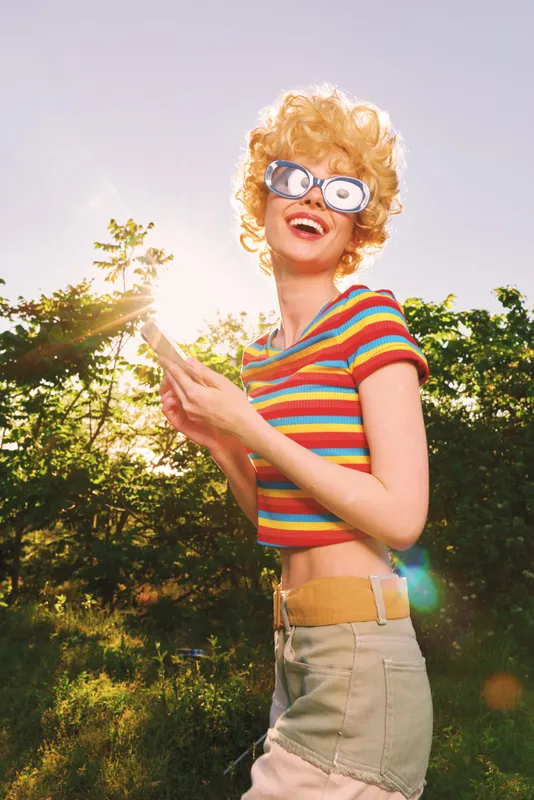 Cheerful woman outdoors, wearing striped crop top and high waisted shorts, sunglasses, sunlit moment with warm retro film colors, carefree vibe against lush greenery background