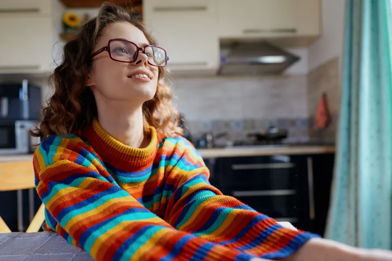 Smiling woman with curly hair wearing a colorful striped sweater in a bright kitchen, relaxed at home, capturing a warm everyday lifestyle moment, soft light