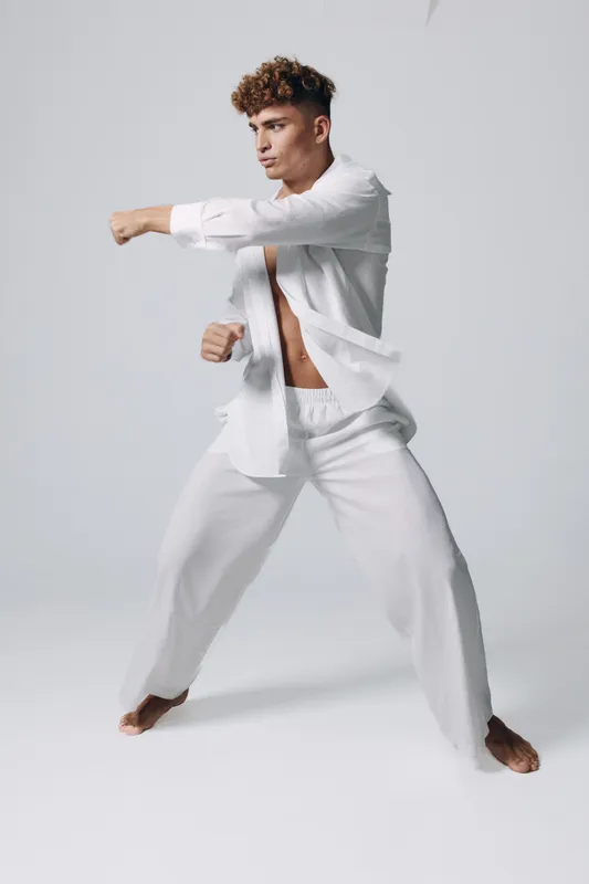 Man in white loose clothing practicing martial arts with focused expression in studio. Barefoot athlete performing punch stance for fitness and self-defense training.