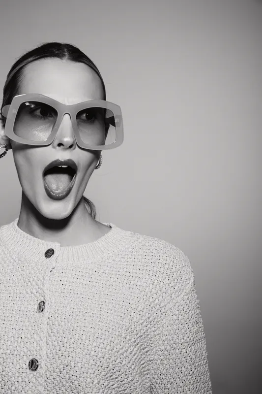 Stylish young white woman with pale skin wearing large sunglasses and knitted sweater shows surprised expression in studio. Concept of fashion, emotion, and modern lifestyle.