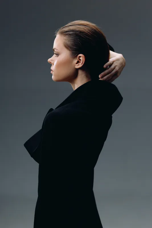 Profile of a thoughtful woman in black clothing posing against gray background with serious expression.