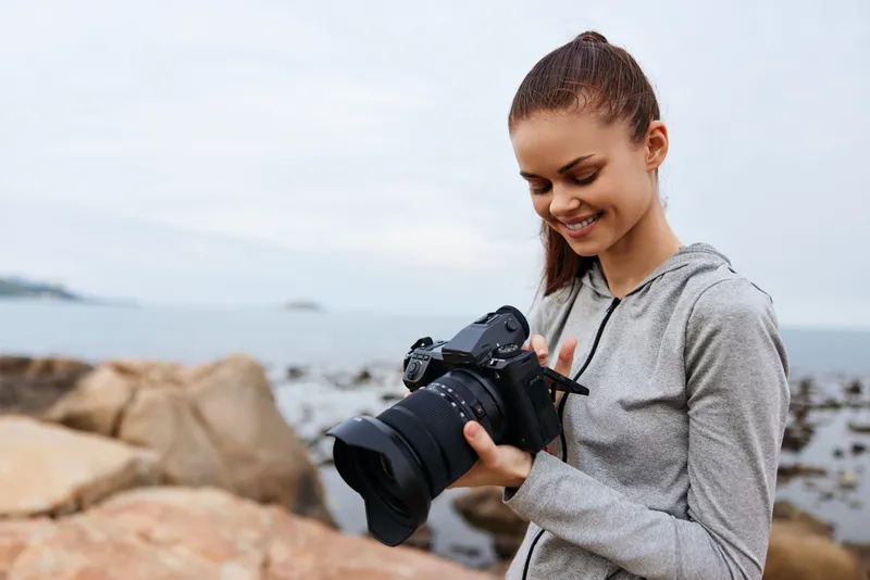 Smiling young woman checking her DSLR camera equipment by the seaside, wearing a casual gray hoodie, joyful expression, with rocky beach and cloudy sky background