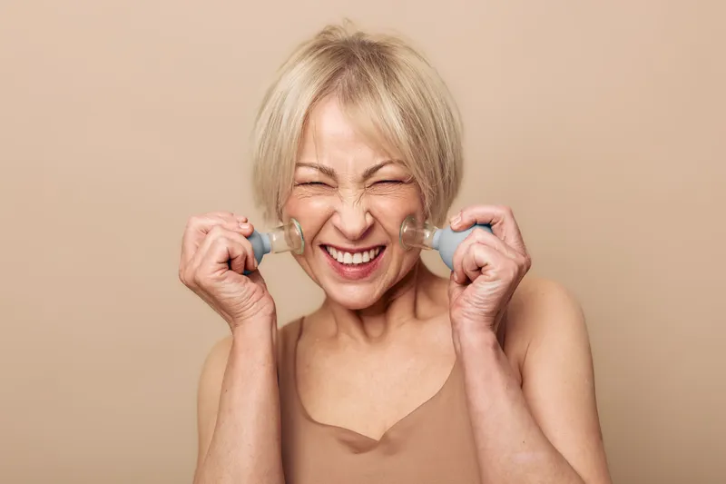 Happy mature woman with short blonde hair uses facial massage cups on cheeks, smiles with closed eyes. Beige studio background, skincare, anti aging, wellness and self care concept.