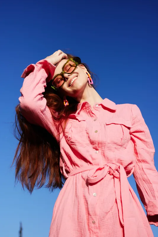 Young woman wearing a pink dress and sunglasses, joyfully posing against a bright blue sky, reflecting a vibrant summer vibe and stylish confidence