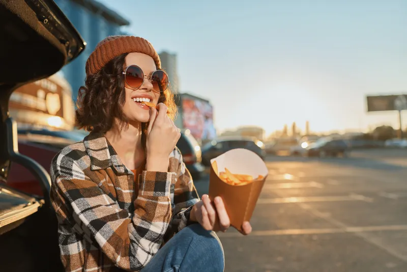 woman fries casual car sunset sunglasses beanie smile capturing a candid street moment of a joyful girl eating fries by an open trunk at golden hour glow, authenticity, mindful living and emotional