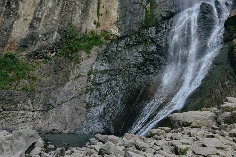 natural waterfall cascading down rocky cliff with clear water flowing into serene pool surrounded by stones and green moss in outdoor environment