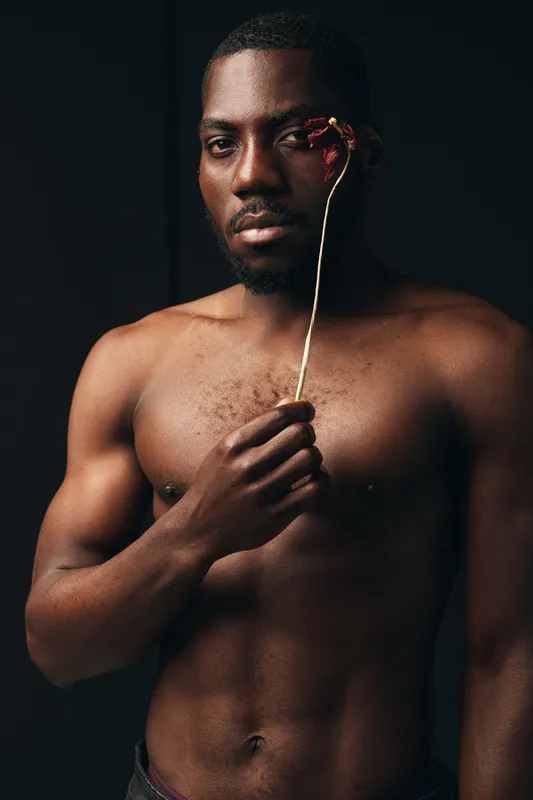 African man shirtless holding dried flower near face in creative conceptual visual style with dark background. Portrait of male model showing emotion and artistic expression in studio.