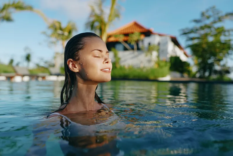 Relaxed woman floats in clear water, eyes closed, soaking sun, amid a tropical resort setting, calm mood, wellness, and outdoor leisure, capturing a peaceful lifestyle moment