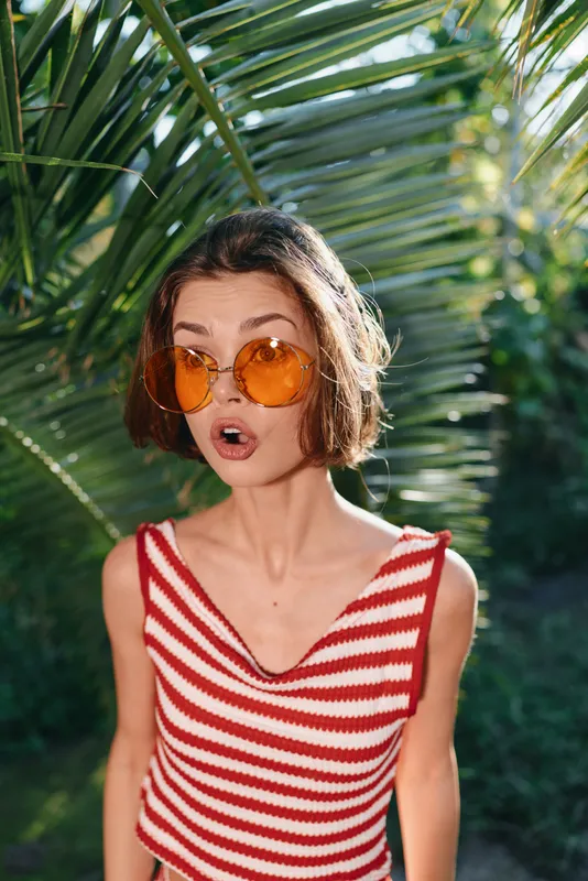 A stylish young woman wearing a red and white striped sleeveless top and orange sunglasses stands outdoors among lush tropical plants, creating a playful summer mood.