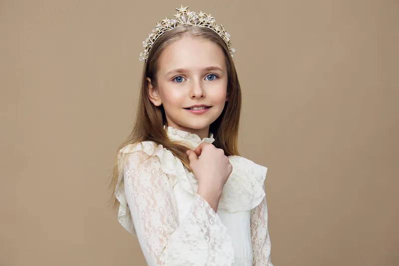 A charming young girl in a lace dress and a crown, smiling softly against a warm beige background, perfect for concepts of childhood beauty and innocence