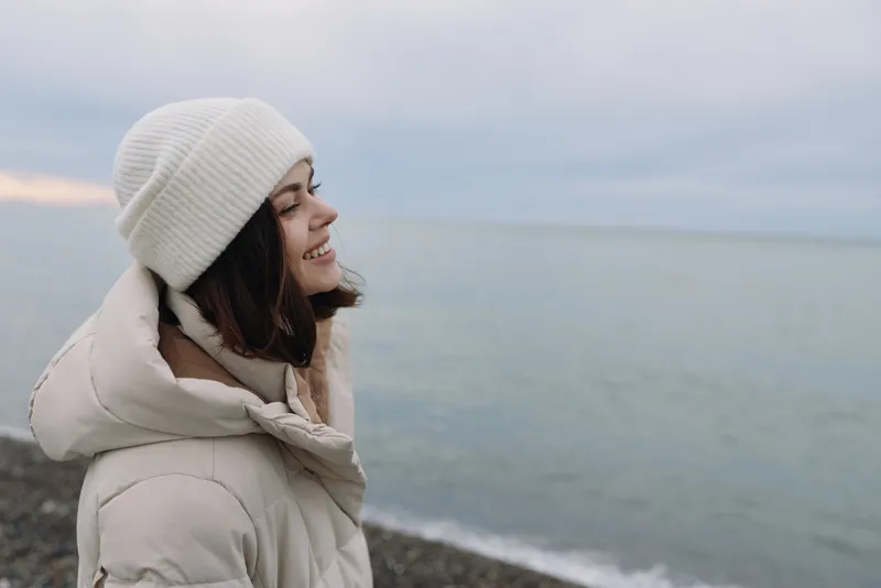 A woman in a light winter coat and knit hat stands beside a calm sea, soft cold light highlights her profile, creating a thoughtful outdoor portrait on the quiet coastline.
