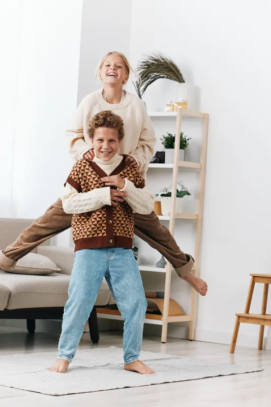 Two joyful children play indoors, one perched on the other's shoulders, laughter fills a bright living room with soft decor, cozy sweater and carefree spirit.
