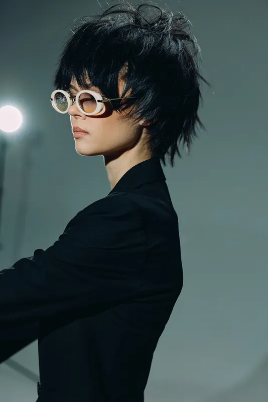 Stylish woman with short black hair wearing round glasses and a black outfit against a muted background.