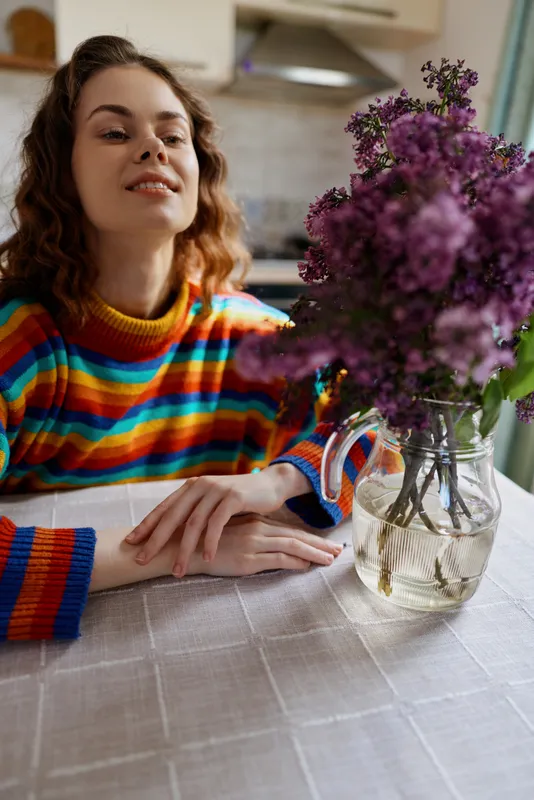 Cheerful woman indoors, relaxed at a dining table, colorful striped sweater, soft natural light, fresh flowers in a vase, calm home moment, candid lifestyle vibe