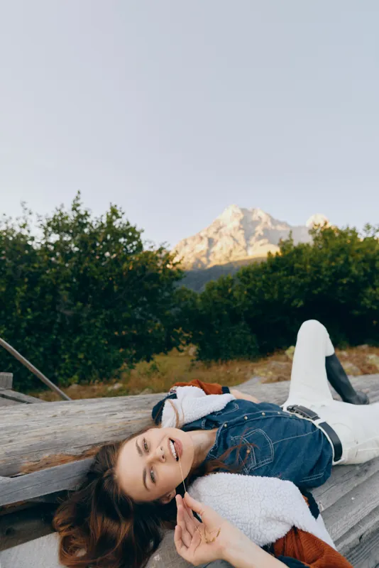 Woman smile outdoor mountain nature relaxed portrait in denim jacket lying on a wooden bench, happy young female enjoying scenic landscape with trees, sunlight and distant peak.