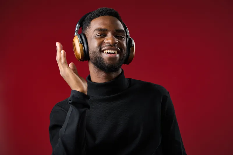 Smiling young man wearing headphones, enjoying music against a vibrant red background, showcasing relaxed emotions and contemporary style