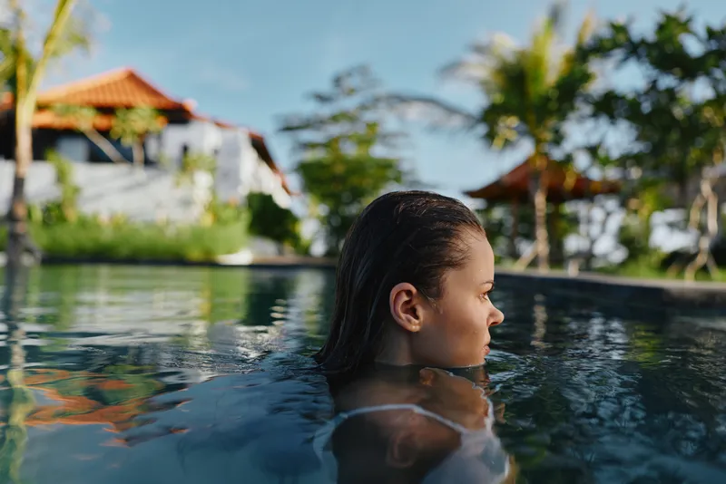 Girl enjoys a sunny poolside moment, wet hair, calm focus, relaxing vacation vibe by a tropical resort, outdoor scene, clear water, bright reflections.