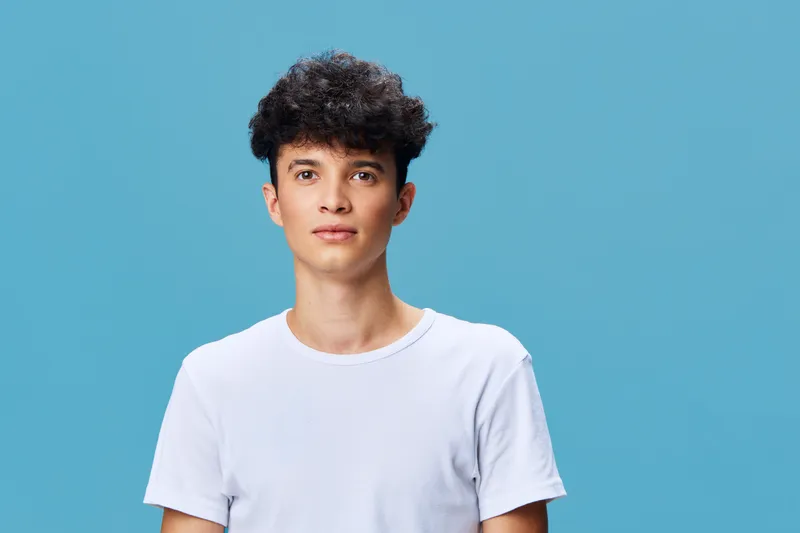 young man with curly hair wearing a plain white t-shirt stands against a solid blue background, showing a neutral expression and casual style