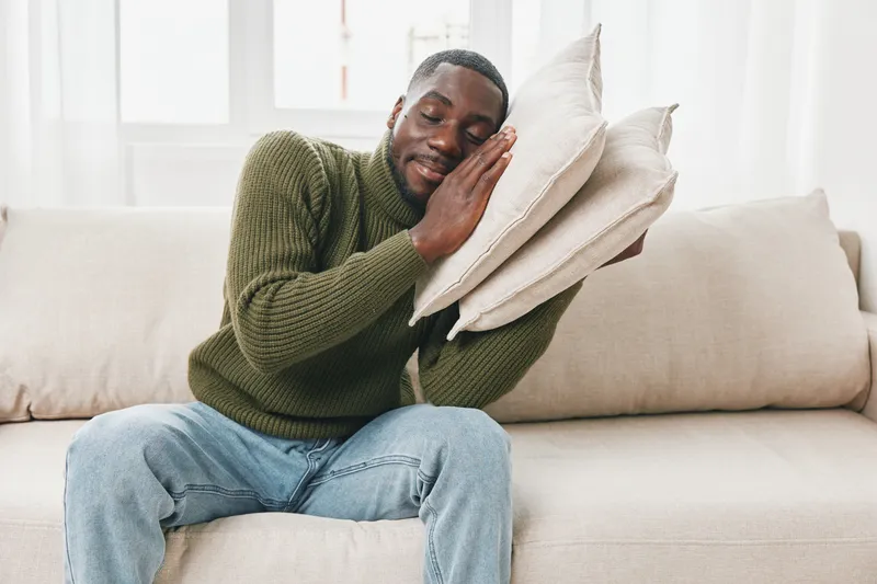 A young Black man in a cozy green sweater hugging decorative pillows on a light colored sofa, portraying a sense of comfort and relaxation in a bright, airy room