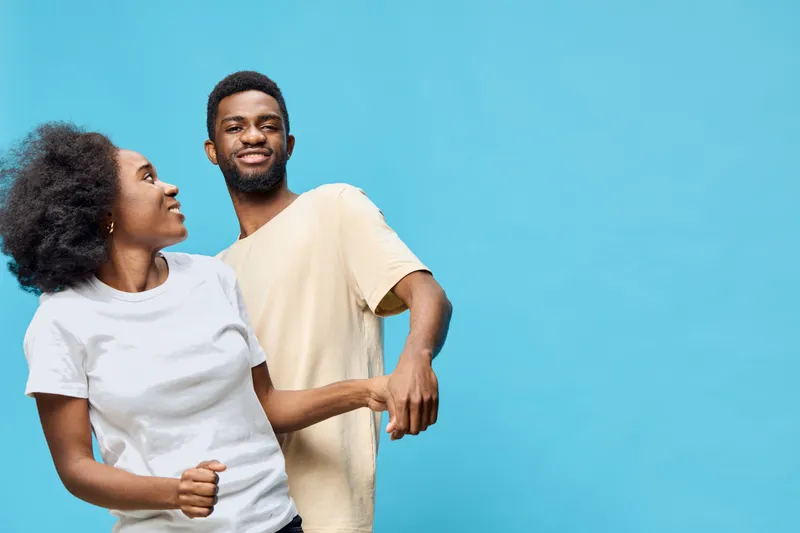 A joyful young couple dancing together against a vibrant blue background, showcasing happiness, connection, and fun in casual attire
