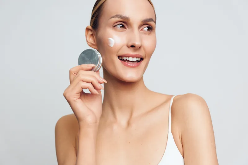 A smiling young woman with fair skin applies cream to her cheek using a round pad in a bright studio, showcasing a simple skincare moment and a sense of routine.
