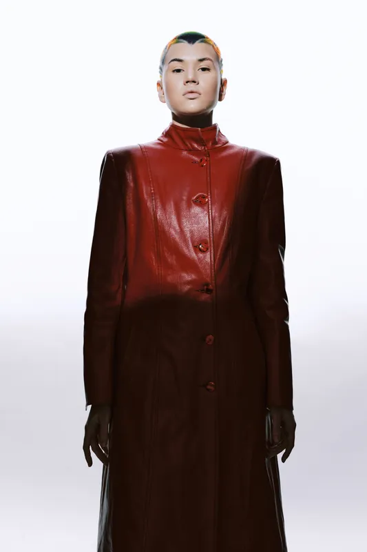 Woman in a long red leather coat stands confidently against a bright white background, showcasing modern fashion with a strong, minimalist style and edgy vibe.