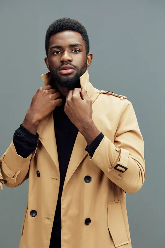 Stylish young African man in a beige trench coat adjusting his collar, showcasing confidence and modern fashion against a muted gray background