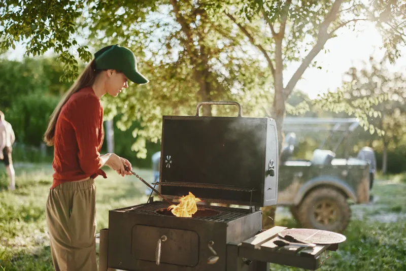 Young woman grilling outdoors, wearing a green cap and red sweater, with flames on the barbecue, surrounded by greenery and warm sunlight in a serene summer setting