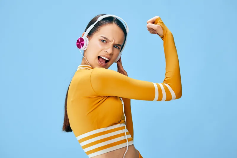 Female athlete in a yellow cropped top flexing arm against a blue background, showcasing fitness, sport and energy. Dynamic pose, headphones on, active lifestyle, strength training and personal