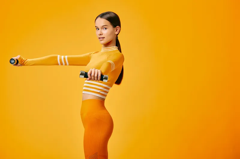 Fit woman in sporty outfit holding dumbbells in a studio workout pose, side profile full body portrait against smooth orange background, conveying strength, balance and active lifestyle.