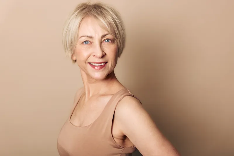 Smiling mature woman with short blonde hair wears beige tank top against neutral background. Studio portrait with soft lighting captures confident and natural beauty concept for aging gracefully.