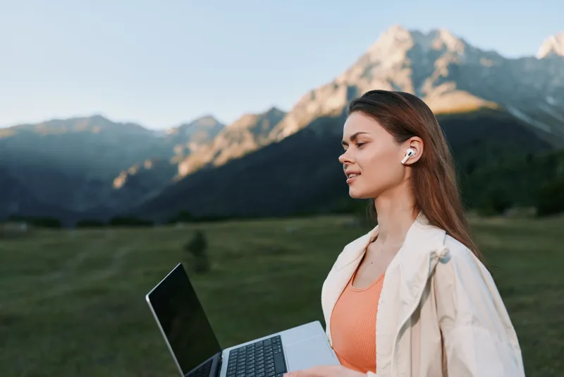 A focused woman sits outdoors with a laptop in a scenic valley, wearing a light jacket and earbud, conveying concentration, calm, and productive atmosphere.