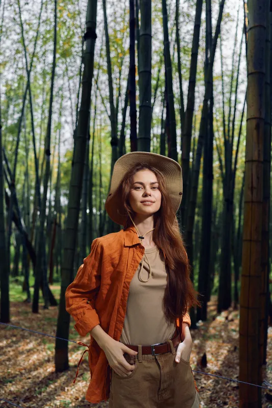 Woman in straw hat portrait standing in bamboo forest, nature scene with longhair, boho outfit and relaxed pose among tall green stalks and dappled sunlight.
