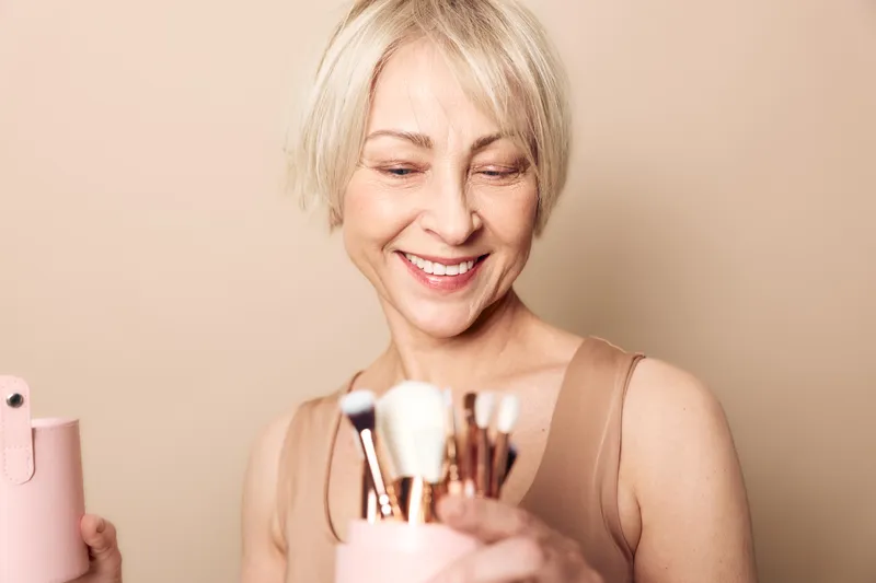 Smiling middle aged woman with short blonde hair holding makeup brushes in a beige studio. Concept of beauty, cosmetics, self care, and joyful lifestyle for mature women.