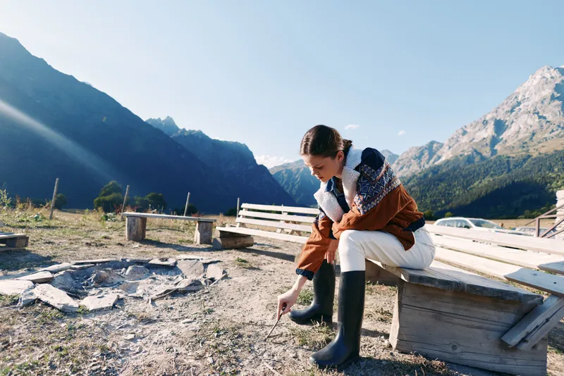 Woman sitting on a wooden bench in autumn mountains, wearing rubber boots and warm jacket in an outdoor lakeside campsite, touching ground and enjoying peaceful nature with clear sky.