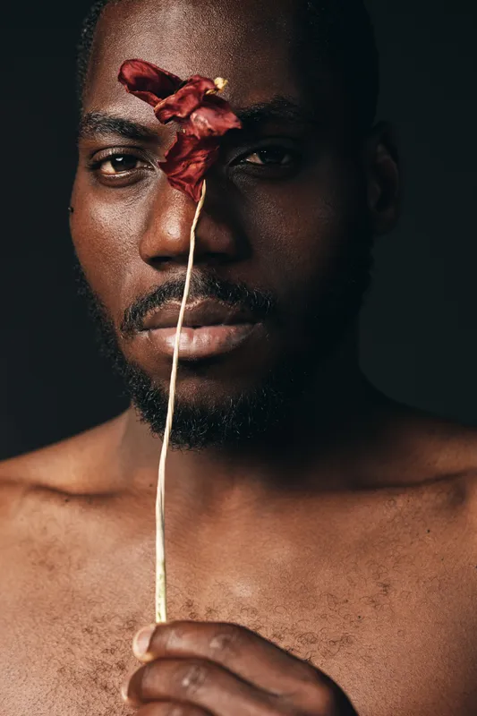 Black man with dark skin holding dried red flower in front of face in creative conceptual visual style. Closeup portrait with serious expression and artistic mood on dark background.