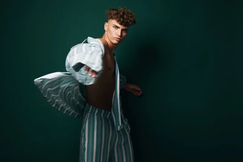 Young man with curly hair wearing striped pajamas posing dynamically against dark background. Fashion, casual style, confident expression and modern look in studio lighting.