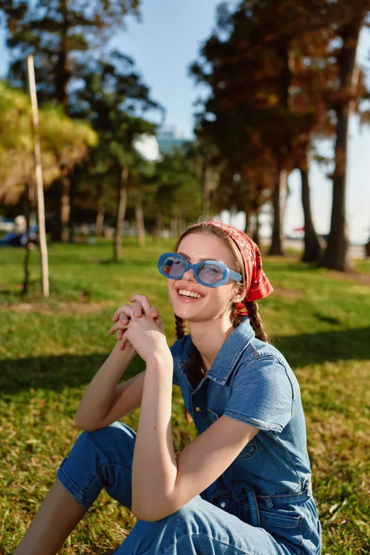 Lifestyle woman in modern boho-western denim street style sitting outdoors with bright blue sunglasses and a red bandana, smiling under natural sunlight with film color effect.