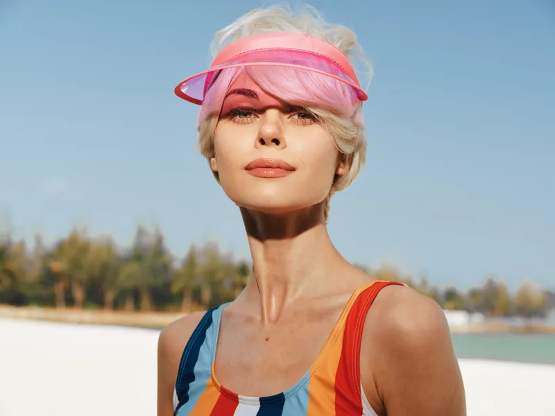 Close up portrait of a stylish woman at the beach wearing a colorful striped swimsuit and pink visor, sunlit skin, serene expression, clear blue sky behind.