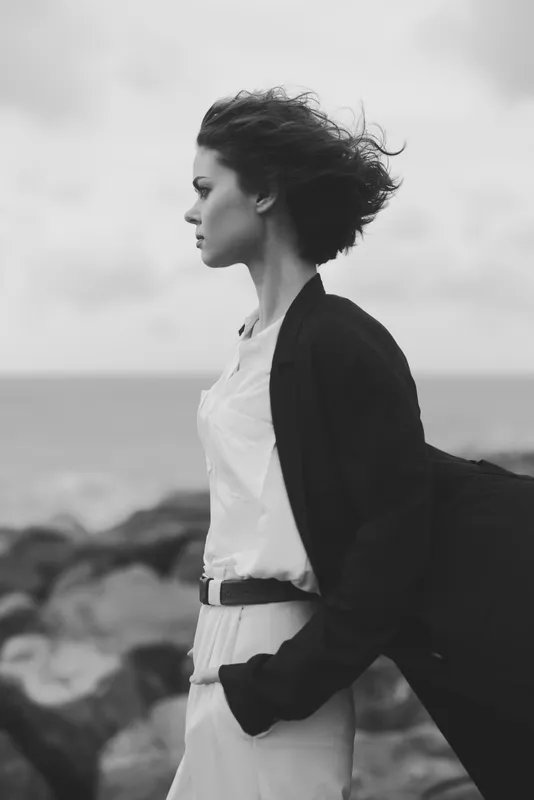 A windy coastal scene shows a stylish woman in a dark blazer and white dress standing on rocky shore, looking toward the horizon, conveying confidence, movement and contemporary fashion editorial